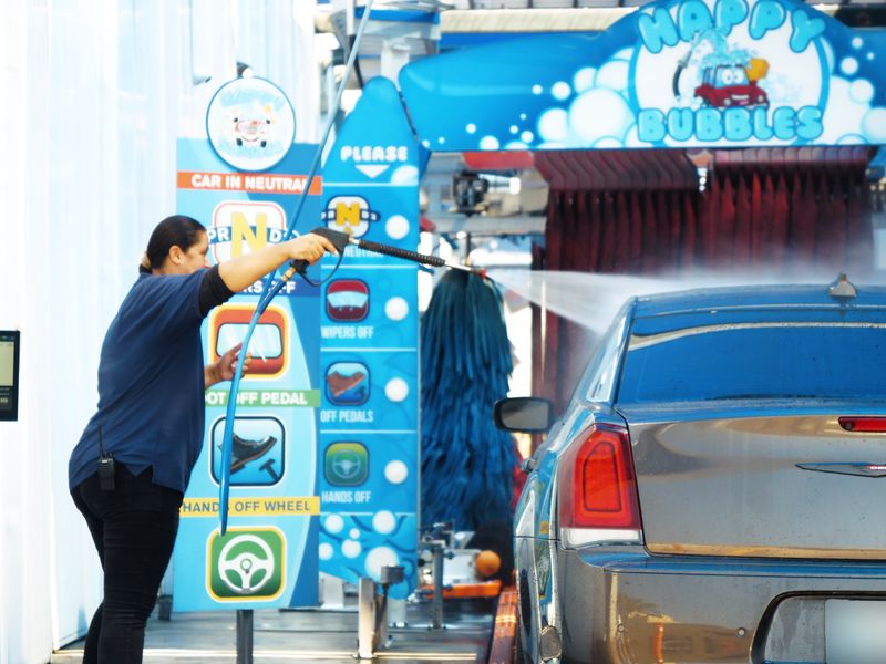 Happy Bubbles team member spraying down a gray sedan at the tunnel entrance under the Happy Bubbles arch